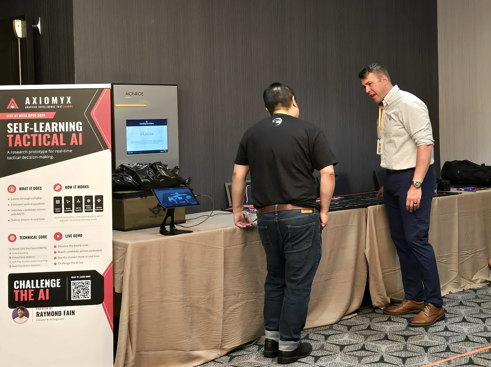 The Axiomyx stand at Nova Open 2026: pull-up banner, demonstration monitor and tablets on the table, with a player and Raymond Fain in conversation beside it.