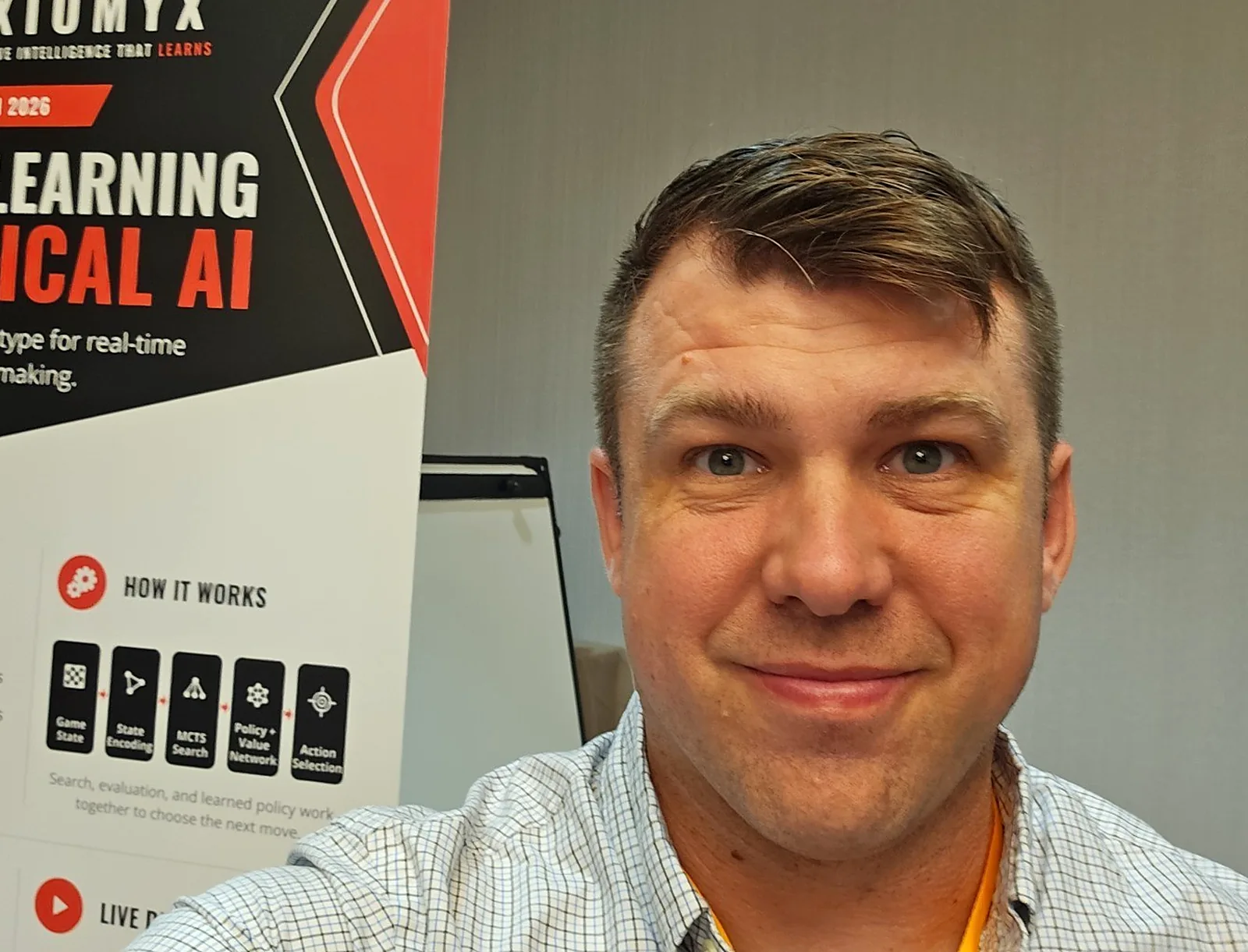 Raymond Fain at the Axiomyx stand at Nova Open 2026, with the banner behind him showing the steps the system works through to choose a move.